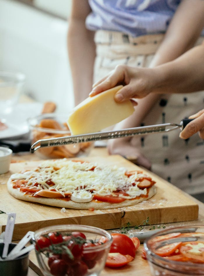 Crop unrecognizable person grating cheese on pizza with champignons and tomatoes while cooking in kitchen
